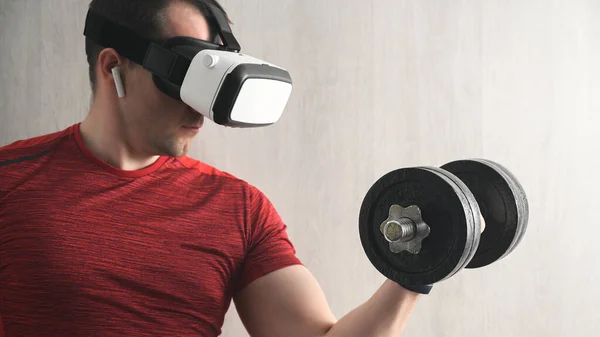 Fitness, sport and technology. Portrait of concentration young adult sport man in a red t-shirt training in home, holding dumbbell with arm, doing exercises for biceps and looking at vr glasses
