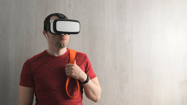 Fitness, workout and VR technology. Portrait Caucasian young adult man in a red t-shirt with an expander on the shoulders, wearing virtual reality glasses. Indoors, copy space