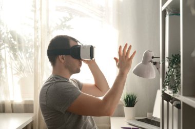 Delikanlı, Covid-19 yüzünden kilitlenme sırasında yurt içi ofisindeki arttırılmış VR kulaklığı kullanıyor. VR kulaklıklı bir erkek. Ev hayatı, pencereden yansıyan doğal ışık.