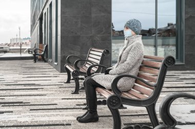 Virüs ve tozu korumak için maske takan gri paltolu beyaz bir kadın, şehir merkezindeki bir bankta oturuyor.