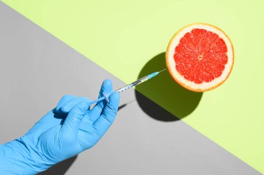 Coronavirus vaccination creative concept. Hand in medical glove holding syringe with vaccine against COVID-19 injecting into grapefruit. Flying objects on pastel green and gray background