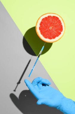 Coronavirus vaccination creative concept. Half grapefruit and a syringe with vaccine against COVID-19, balancing on a hand in a medical glove. Minimalistic creative image on pastel green background