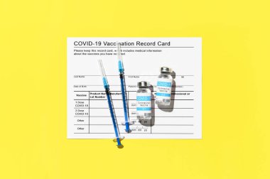Coronavirus vaccination concept. Top view of vaccination record card, syringes, and two vials of the COVID-19 vaccine for two-stage injection. Bright yellow background