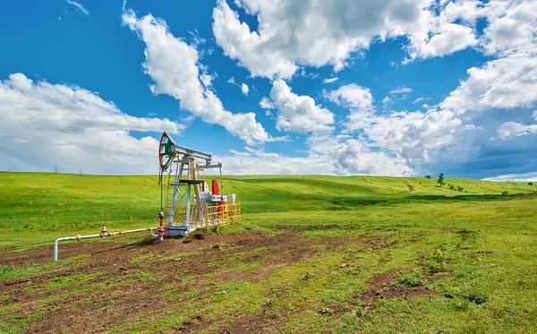 Unidad de bombeo para bombear aceite en un prado verde contra un cielo azul con nubes en verano —  Fotos de Stock