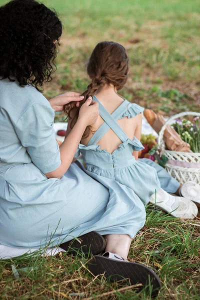 青いドレスの母親は娘の髪を編んだ 女性は少女を編組にしている 娘のための美しい髪型の概念 公園のピクニックで幸せな家族 — ストック写真