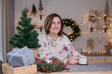 Masada oturan ve Noel süslemeli odada sıcak çikolata içen bir kadın. Noel zamanı mutlu bir anne. Mutfakta Noel süsü var. Evde yeni yıl..