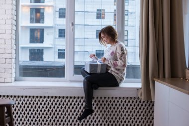 Panoramik bir pencerenin pencere eşiğinde oturan genç bir kadın. Evde hediyesiyle. Kadın pencereye oturmuş elinde tatil hediyesi taşıyor.