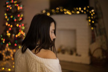 Beyaz kazaklı bir kadın Noel ağacı ve şöminenin önünde oturuyor. Noel arifesinde siyah saçlı güzel bir kadın. Sıcak tatiller. Aydınlatılmış bokeh