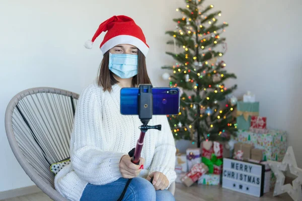 Woman in medical mask near christmas tree having a video call on phone, enjoy Christmas holidays at home with Christmas tree. Selfie in medical protective mask and santa hat