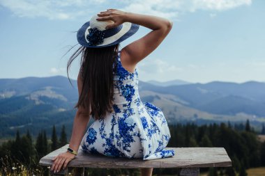 Şık elbiseli genç bir kadının, dağlardaki bankta otururken nefes kesen manzarasına hayran olduğu bir sahne. Rüzgâr yüzünden şapkasını kafasında tutmak için.