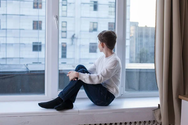 The boy sits on the windowsill and looks out the window — Stock Photo, Image