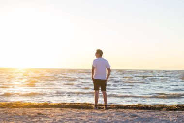 Beyaz gömlekli ve kahverengi şortlu bir adam gün doğumunda boş sahilde duruyor..
