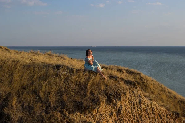 A girl in a long dress sits on a cliff by the sea