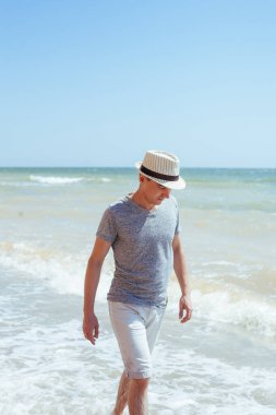 Gri gömlekli, beyaz şortlu ve hasır şapkalı bir adam sahilde yürüyor.