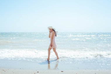 Pembe elbiseli ve hasır şapkalı bir kadın sahilde yürüyor.