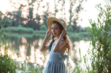 Mavi elbiseli ve hasır şapkalı bir kız gölün kıyısında duruyor.