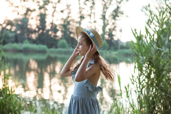 Una chica en vestido azul y sombrero de paja se encuentra en la orilla del lago — Foto de Stock