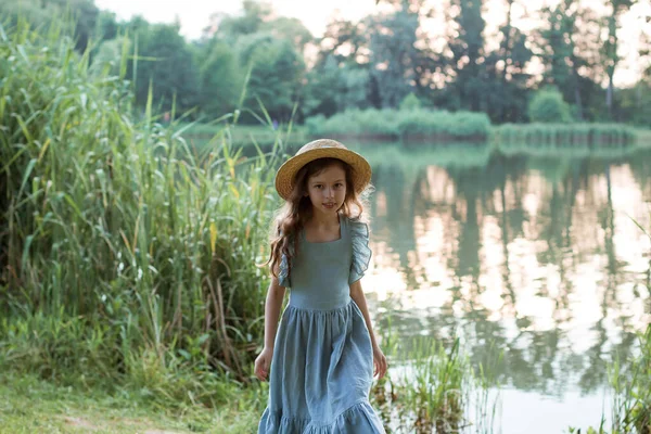 Dziewczyna w niebieskiej sukience i słomkowym kapeluszu stoi na brzegu jeziora — Zdjęcie stockowe