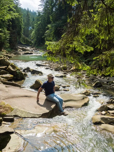 Мужчина в соломенной шляпе, футболке и джинсах сидит у горной реки — стоковое фото