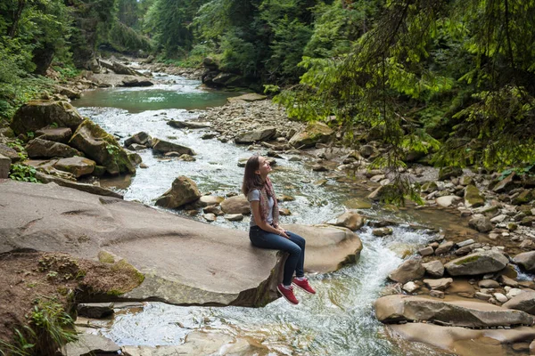 Chica en una camiseta blanca y vaqueros azules sentado cerca de un río de montaña —  Fotos de Stock