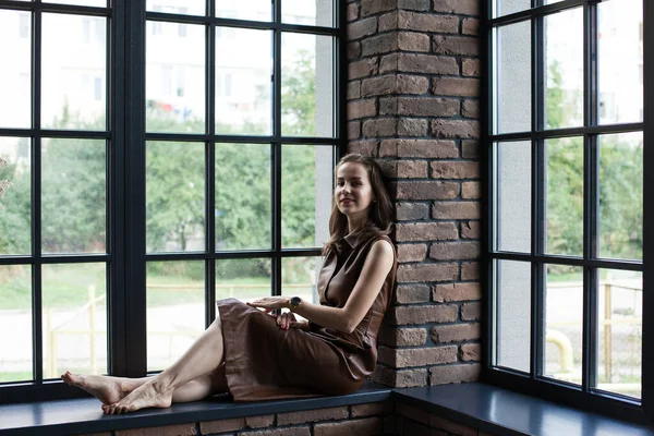 Schöne junge Frau in braunem Lederkleid sitzt barfuß auf der Fensterbank — Stockfoto