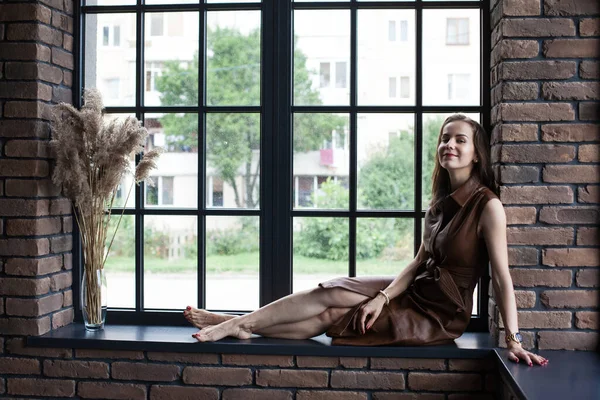 Belle jeune femme en robe en cuir marron assise sur le rebord de la fenêtre pieds nus — Photo