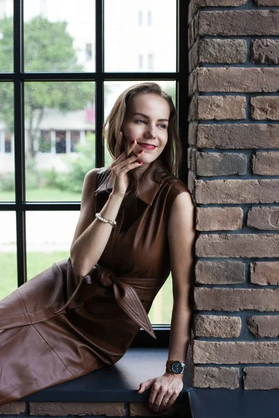 Schöne junge Frau im braunen Lederkleid sitzt auf der Fensterbank — Stockfoto