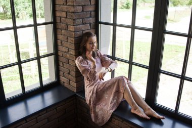 Pembe elbiseli güzel bir kadın pencerenin kenarında çıplak ayakla oturuyor.