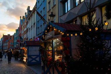 Gdansk, Polonya, 27 Kasım 2025: Mallarla dolu tezgahlar cadde boyunca oturur. Güneş batarken insanlar etrafta dolaşır. Noel pazarında ışıklar parlıyor..