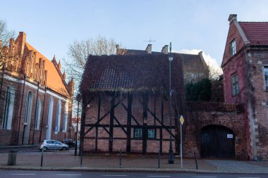 Bir caddenin yanında sazdan çatılı tarihi bir bina duruyor. Etrafını saran diğer yapılar ve yakınlarda ağaçlar görülebilir..