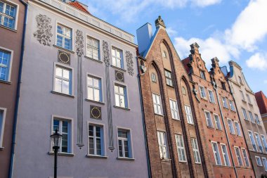 Polonya 'nın Gdansk kentindeki bir caddede birçok tarihi bina yan yana duruyor. Gökyüzünde bulutlar süzülürken güneş ön cephelerde parlıyor..