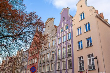 Polonya 'nın Gdansk şehrinde gündüz saatlerinde renkli binalar ve ağaçlarla dolu bir sokakta yürümek. Gökyüzü birkaç bulutla mavidir.