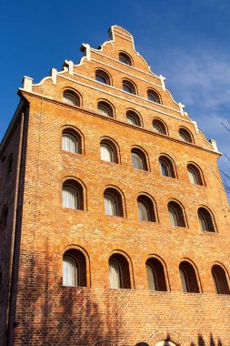 Mavi bir gökyüzünün altında birkaç kemer ve penceresi olan tuğla bir bina ayakta duruyor. Yapı, bir şehrin tarihi ve mimarisinin bir karışımını gösteriyor. Gdansk, Polonya