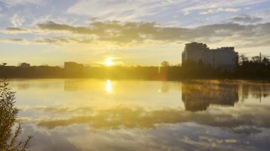 Güneş gölün üzerinden doğar ve altın yansımalar yaratır. Yakınlarda ağaçlarla ve bulutlu gökyüzüyle çevrili bir bina suyun yanında duruyor. Ivano-Frankivsk, Ukrayna