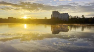 Güneş gölün üzerinden doğar ve altın yansımalar yaratır. Yakınlarda ağaçlarla ve bulutlu gökyüzüyle çevrili bir bina suyun yanında duruyor. Ivano-Frankivsk, Ukrayna