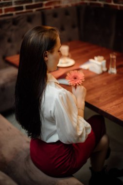 Beyaz gömlekli ve siyah saçlı bir kız, bir masada oturuyor, arkası dönük, elinde bir çiçek var.