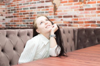 Çok güzel gülümseyen bir kız bir kafedeki kanepede oturuyor ve bir şeyin hayalini kuruyor.