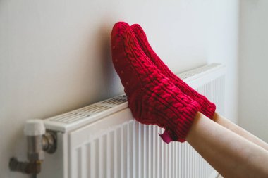 Sıcak, rahat çorap ören genç bir kadın kaloriferin yanında bacaklarını ısıtıyor.