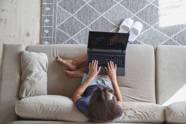 Kanepede oturan ve evinde dizüstü bilgisayar kullanan mutlu genç bir kadın..