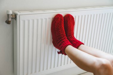 Sıcak, rahat çorap ören genç bir kadın kaloriferin yanında bacaklarını ısıtıyor.