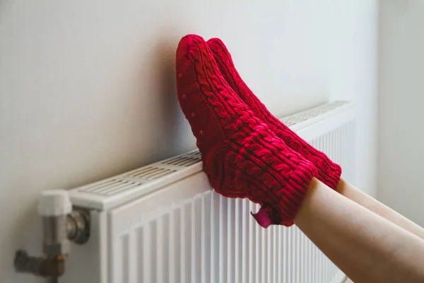 Sıcak, rahat çorap ören genç bir kadın kaloriferin yanında bacaklarını ısıtıyor.