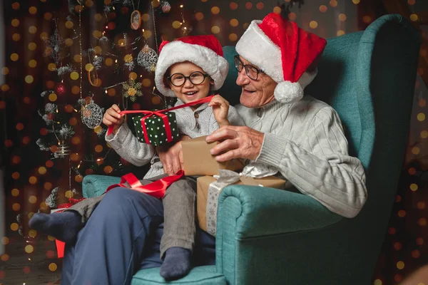 年事已高的祖父头戴圣诞礼帽 在一棵圣诞树的背景下送出并打开圣诞礼物 — 图库照片