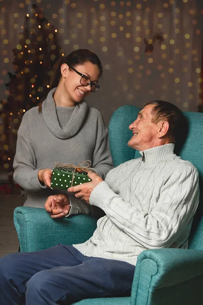 一位年迈的父亲和他年幼的女儿在一棵圣诞树的背景下送给并打开了一份圣诞礼物 — 图库照片