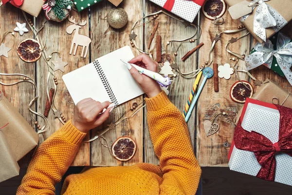 Woman hands with a pen and notepad writing letter to santa claus or christmas shopping list, presents on table with christmas gifts and decorations