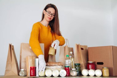 Tıbbi eldiven takan genç bir kadın, yiyecek bağışlarını kese kağıdına dolduruyor.