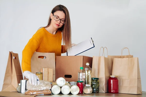 Tıbbi eldivenlerle gönüllü olan genç bir kadın kağıt torbaya yiyecek doldurup yazıyor.