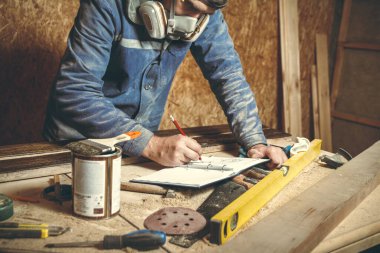 Erkek marangoz kendi stüdyosunda ahşapla çalışıyor ve kalemle çizimler yapıyor.