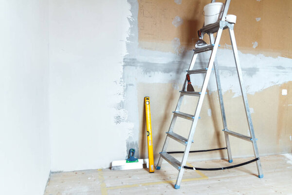 A stepladder with painting tools in a room in a house or apartment. Preparing for putty on the wall or painting. Home repair or renovation concept.