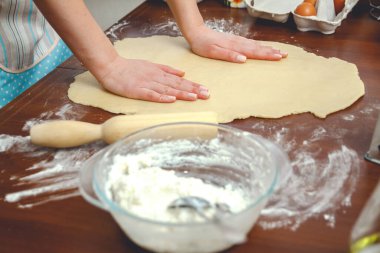 Mutfakta yemek yapan genç bir kadın, masada pişirmek için hamuru oklavayla yuvarlıyor.