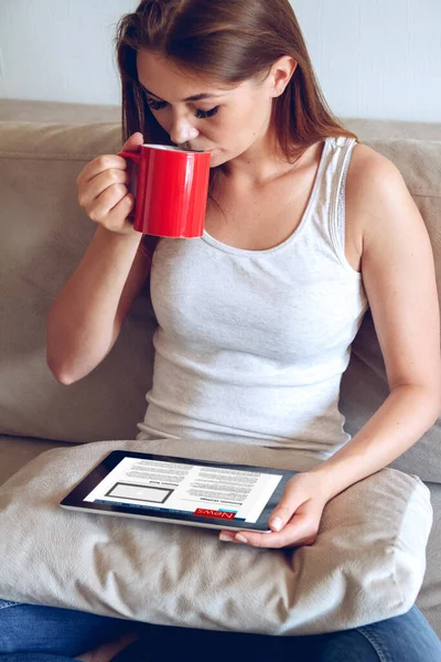 Kot Pantolonlu Bir Kadın Kanepeye Oturmuş Tablet Kullanıyor Kırmızı Bardaktan — Stok fotoğraf